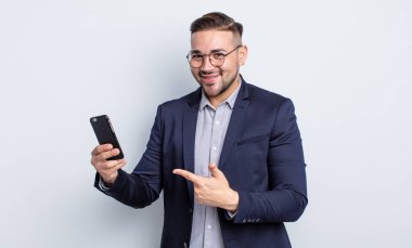 Genç yakışıklı adam neşeyle gülümsüyor, mutlu hissediyor ve ek iş ve akıllı telefon kavramına işaret ediyor.