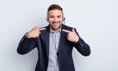 Genç ve yakışıklı bir adam kendi gülüşünü işaret ederek kendine güvenerek gülümsüyor. Tele pazarlamacı kavramı