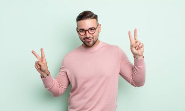 Genç yakışıklı adam gülümsüyor ve mutlu görünüyor, arkadaş canlısı ve memnun, işaret eden zafer ya da barış iki elle