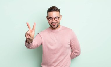 Genç yakışıklı adam gülümsüyor ve mutlu görünüyor, kaygısız ve olumlu, zafer veya barış işareti yapıyor.