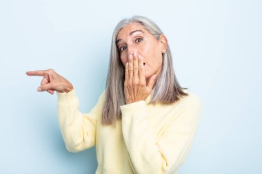Orta yaşlı beyaz saçlı kadın kendini mutlu, şaşkın ve şaşırmış hissediyor. Ağzını eliyle kapatıyor ve yanal fotokopi alanını işaret ediyor.