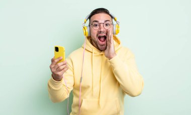 Genç yakışıklı adam mutlu hissediyor, elleri ağzının kenarında bağırıyor. kulaklıklar ve akıllı telefon konsepti