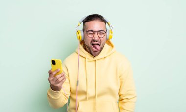 Genç, yakışıklı, neşeli ve asi tavırlı, şaka yapan ve dilini dışarı çıkaran bir adam. kulaklıklar ve akıllı telefon konsepti