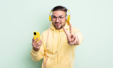 Genç yakışıklı adam gülümsüyor ve arkadaş canlısı görünüyor, iki numarayı gösteriyor. kulaklıklar ve akıllı telefon konsepti