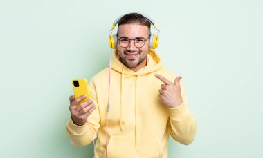 Genç ve yakışıklı bir adam kendi gülüşünü işaret ederek kendine güvenerek gülümsüyor. kulaklıklar ve akıllı telefon konsepti