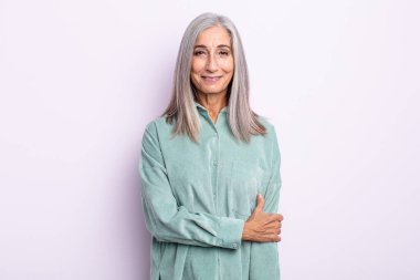 Orta yaşlı gri saçlı kadın utangaç ve neşeli gülüyor, arkadaş canlısı, olumlu ama kendine güveni olmayan bir tavırla