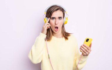 Genç, güzel bir kadın şaşırmış görünüyor, yeni bir düşünce, fikir ya da konsept fark ediyor. kulaklıklar ve telefon konsepti