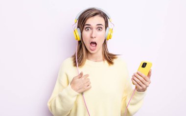 Şok olmuş ve ağzı açık, kendini gösteren genç ve güzel bir kadın. kulaklıklar ve telefon konsepti