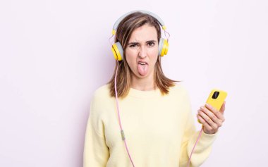 Genç ve güzel bir kadın iğreniyor, sinirleniyor ve dilini çıkarıyor. kulaklıklar ve telefon konsepti