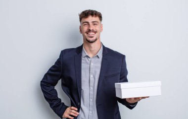 Genç yakışıklı adam mutlu bir şekilde gülümsüyor, eli kalçasında ve kendinden emin. beyaz kutu paketi