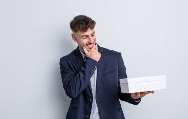 Genç ve yakışıklı bir adam mutlu ve kendinden emin bir ifadeyle gülümsüyor, elleri çenesinde. beyaz kutu paketi