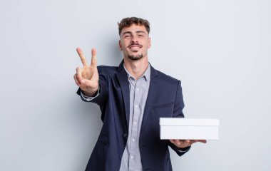 Genç yakışıklı adam gülümsüyor ve mutlu görünüyor, zafer ya da barış işareti yapıyor. beyaz kutu paketi