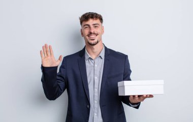 Genç yakışıklı adam mutlu bir şekilde gülümsüyor, el sallıyor, sizi selamlıyor ve selamlıyor. beyaz kutu paketi