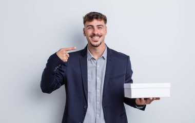 Genç ve yakışıklı bir adam kendi gülüşünü işaret ederek kendine güvenerek gülümsüyor. beyaz kutu paketi