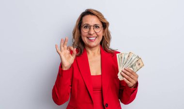 Orta yaşlı güzel bir kadın kendini mutlu hissediyor. Onayını iyi bir jestle gösteriyor. Dolar banknotları konsepti