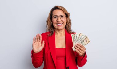 Güzel orta yaşlı bir kadın mutlu bir şekilde gülümsüyor, el sallıyor, sizi selamlıyor ve selamlıyor. Dolar banknotları konsepti