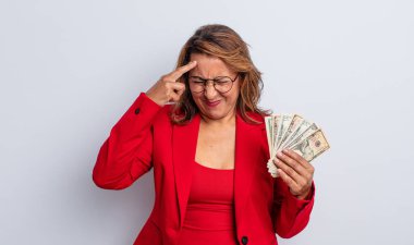 Oldukça orta yaşlı bir kadın mutsuz ve stresli görünüyor, intihar jesti silah işareti yapıyor. Dolar banknotları konsepti