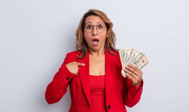 Oldukça orta yaşlı bir kadın şok olmuş ve ağzı açık, kendini işaret ederek şaşırmış görünüyor. Dolar banknotları konsepti