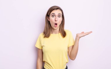 Genç ve güzel bir kız şaşırmış ve şok olmuş görünüyor, çenesi düşmüş, bir objeyi elinde açık bir elle tutuyor.