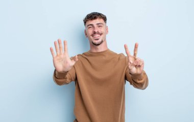 Genç yakışıklı adam gülümsüyor ve dost canlısı görünüyor, 7 ya da 7 numarayı gösteriyor, eli önde, geriye doğru sayıyor.