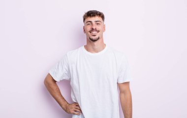 Genç yakışıklı adam mutlu bir şekilde gülümsüyor, eli kalçasında ve kendinden emin, olumlu, gururlu ve arkadaş canlısı.