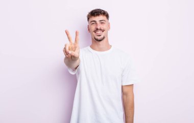 Genç yakışıklı adam gülümsüyor ve mutlu görünüyor, kaygısız ve olumlu, zafer veya barış işareti yapıyor.