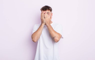 Genç yakışıklı adam elleriyle yüzünü kapatıyor, parmaklarının arasından şaşkın bir ifadeyle yan tarafa bakıyor.