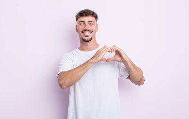 Genç yakışıklı adam gülümsüyor ve mutlu, sevimli, romantik ve aşık, iki eliyle kalbi şekillendiriyor.