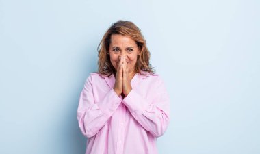 Orta yaşlı bir kadın mutlu, neşeli, şanslı ve şaşkın görünüyor. İki eliyle ağzını kapatıyor.