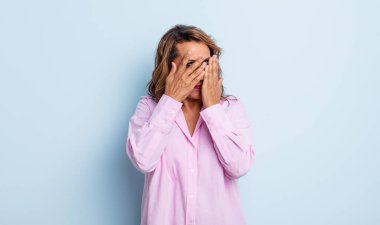 Orta yaşlı kadın elleriyle yüzünü kapatıyor, parmaklarının arasından şaşkın bir ifadeyle yan tarafa bakıyor.