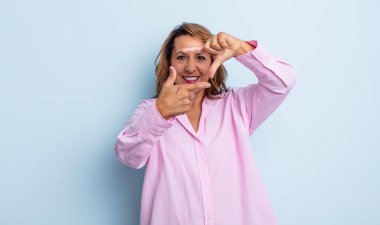 Orta yaşlı bir kadın mutlu, arkadaş canlısı ve pozitif hissediyor, gülümsüyor ve elleriyle bir portre veya fotoğraf çerçevesi yapıyor.