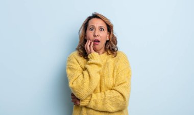 Orta yaşlı bir kadın şok ve inançsızlık içinde, elleri yanağında ve kolları çaprazlanmış, sersemlemiş ve şaşırmış hissediyor.