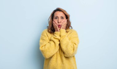 Orta yaşlı kadın şok olmuş ve korkmuş hissediyor, ağzı açık ve elleri yanaklarında korkmuş görünüyor.