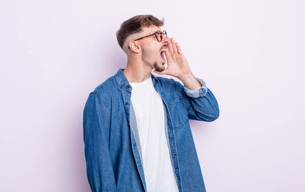 young handsome man yelling loudly and angrily to copy space on the side, with hand next to mouth