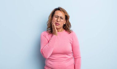 Orta yaşlı bir kadın yanağını tutuyor ve ağrılı diş ağrısı çekiyor. Hasta, sefil ve mutsuz hissediyor. Dişçi arıyor.