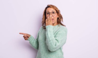 Orta yaşlı kadın kendini mutlu, şaşkın ve şaşırmış hissediyor. Ağzını eliyle kapatıyor ve yanal fotokopi alanını işaret ediyor.