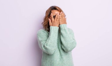 Orta yaşlı kadın elleriyle yüzünü kapatıyor, parmaklarının arasından şaşkın bir ifadeyle yan tarafa bakıyor.