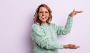 Orta yaşlı kadın gülümsüyor, mutlu, pozitif ve tatmin olmuş hissediyor, fotokopi alanında nesne veya konsept tutuyor veya gösteriyor