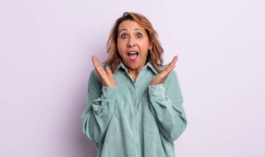 Orta yaşlı bir kadın mutlu ve heyecanlı görünüyor. Beklenmedik bir sürprizle şoke olmuş. İki eli yüzü açık.