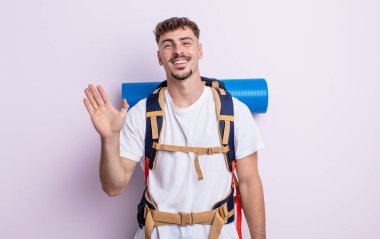 Genç yakışıklı adam mutlu bir şekilde gülümsüyor, el sallıyor, sizi selamlıyor ve selamlıyor. Yürüyüş konsepti