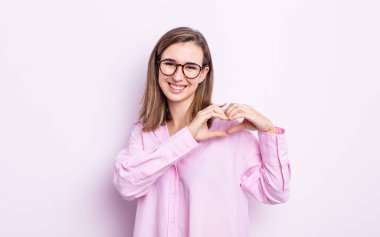 Genç ve güzel bir kız gülümsüyor ve mutlu, sevimli, romantik ve aşık, iki eliyle kalbi şekillendiriyor.