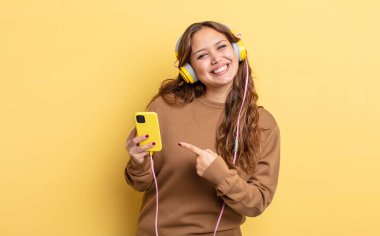 İspanyol güzel kadın neşeyle gülümsüyor, mutlu hissediyor ve bir tarafı işaret ediyor. kulaklıklar ve akıllı telefon konsepti