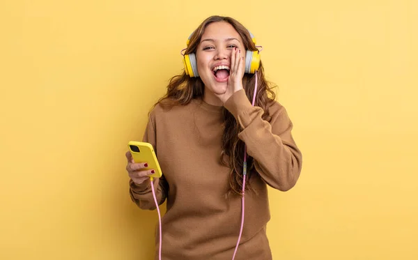 ヒスパニック系のかなりの女性は 手の口の横に大きな声を出す幸せを感じている ヘッドフォンとスマートフォンのコンセプト — ストック写真