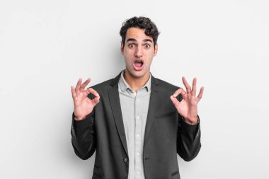 Genç işadamı şok olmuş, şaşırmış ve şaşırmış hissediyor. İki eliyle de onay işareti yapıyor.