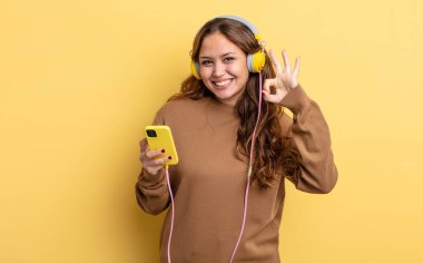 İspanyol güzel kadın kendini mutlu hissediyor, iyi bir jestle onaylıyor. kulaklıklar ve akıllı telefon konsepti
