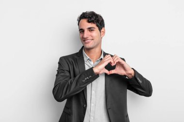 Genç işadamı gülümsüyor ve mutlu, sevimli, romantik ve aşık, iki eliyle kalbi şekillendiriyor.