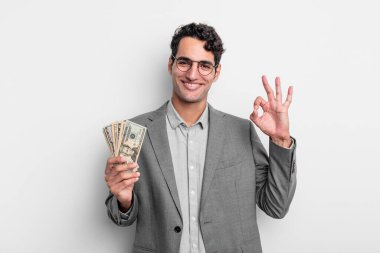 İspanyol yakışıklı adam kendini mutlu hissediyor, iyi bir jestle onaylıyor. iş ve dolar banknotları kavramı