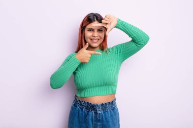 Genç İspanyol kadın mutlu, arkadaş canlısı ve pozitif hissediyor, gülümsüyor ve elleriyle bir portre veya fotoğraf çerçevesi yapıyor.