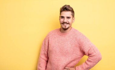 Genç yakışıklı adam mutlu bir şekilde gülümsüyor, eli kalçasında ve kendinden emin, olumlu, gururlu ve arkadaş canlısı bir tavırla. Garip bıyık konsepti