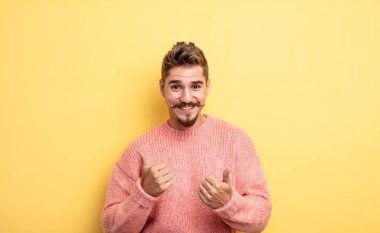 Genç ve yakışıklı adam neşeyle gülümsüyor ve mutlu görünüyor, kaygısız ve olumlu hissediyor iki başparmağıyla. Garip bıyık konsepti